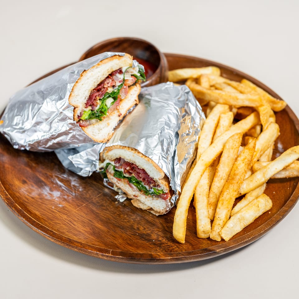 Two foil-wrapped sandwiches cut in half with seasoned french fries and a small bowl of sauce on a wooden plate.