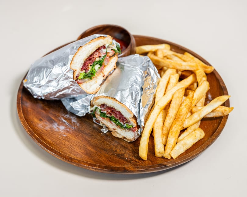 Two foil-wrapped sandwiches cut in half with seasoned french fries and a small bowl of sauce on a wooden plate.