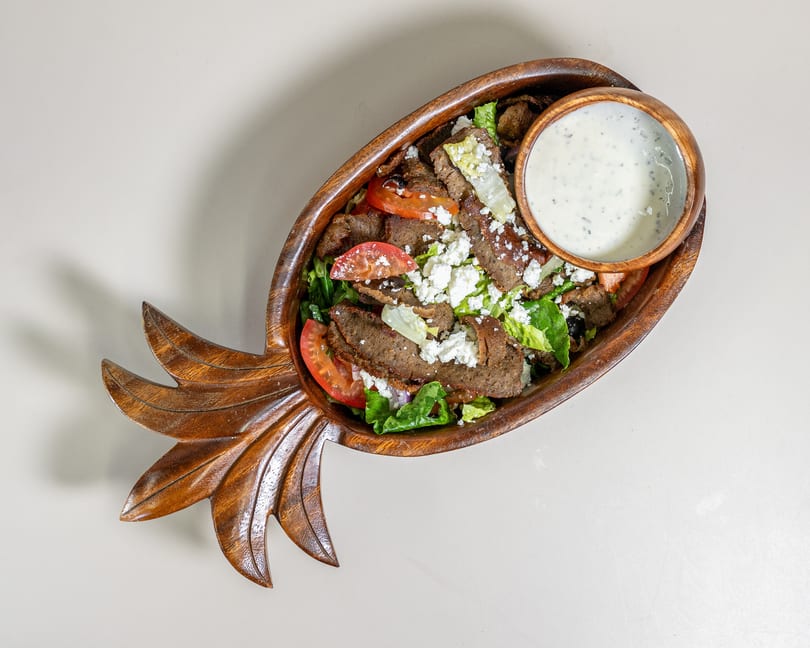 Gyro meat salad with feta, tomatoes, and lettuce in a pineapple-shaped wooden bowl with dressing.