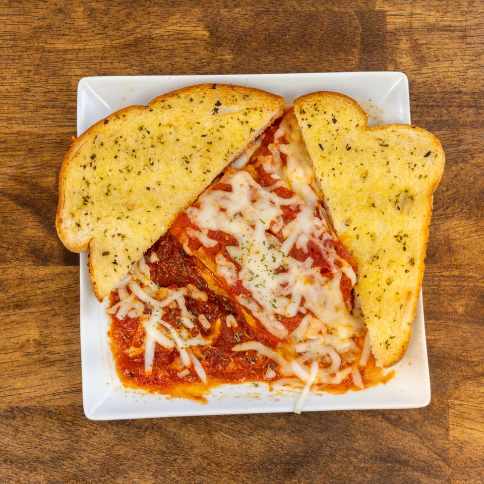 Cheesy lasagna with red sauce and two slices of garlic bread on a white plate.