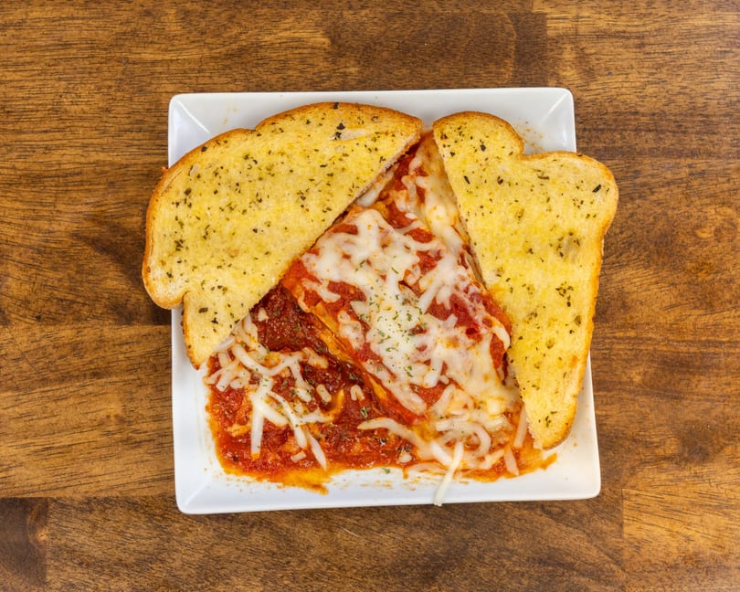 Cheesy lasagna with red sauce and two slices of garlic bread on a white plate.