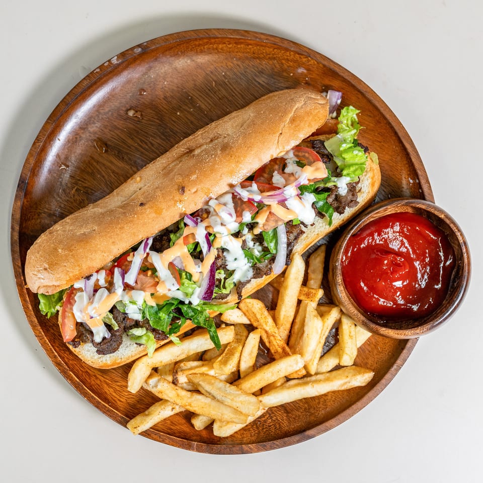 Long sub sandwich with meat, lettuce, tomato, onion, and sauce, served with fries and ketchup on a wooden platter.