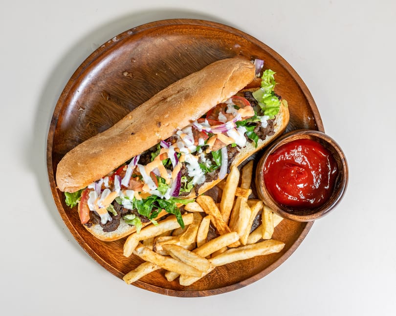 Long sub sandwich with meat, lettuce, tomato, onion, and sauce, served with fries and ketchup on a wooden platter.